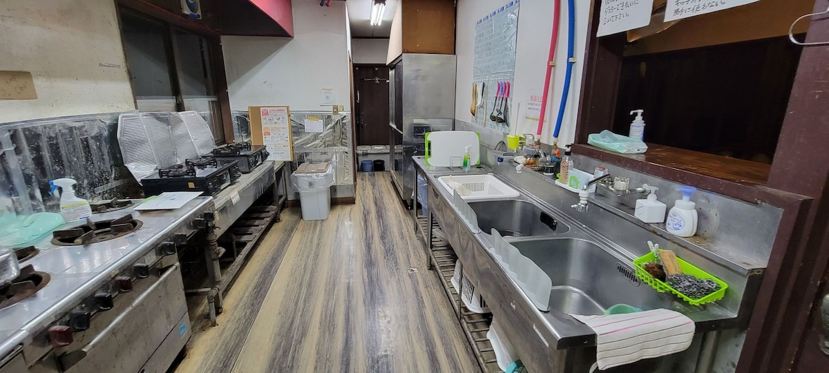 A spacious kitchen is visible, featuring multiple cooking stations with stovetops and a large stainless steel sink area. Countertops are equipped with utensils and kitchenware. Fluorescent lighting provides ample illumination, while wooden flooring adds warmth to the environment.