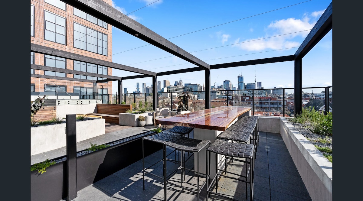 A spacious rooftop terrace showcases an outdoor dining area with a long table and black wicker stools. Surrounding greenery adds a touch of nature, while city skyline views are visible in the background under a clear blue sky.