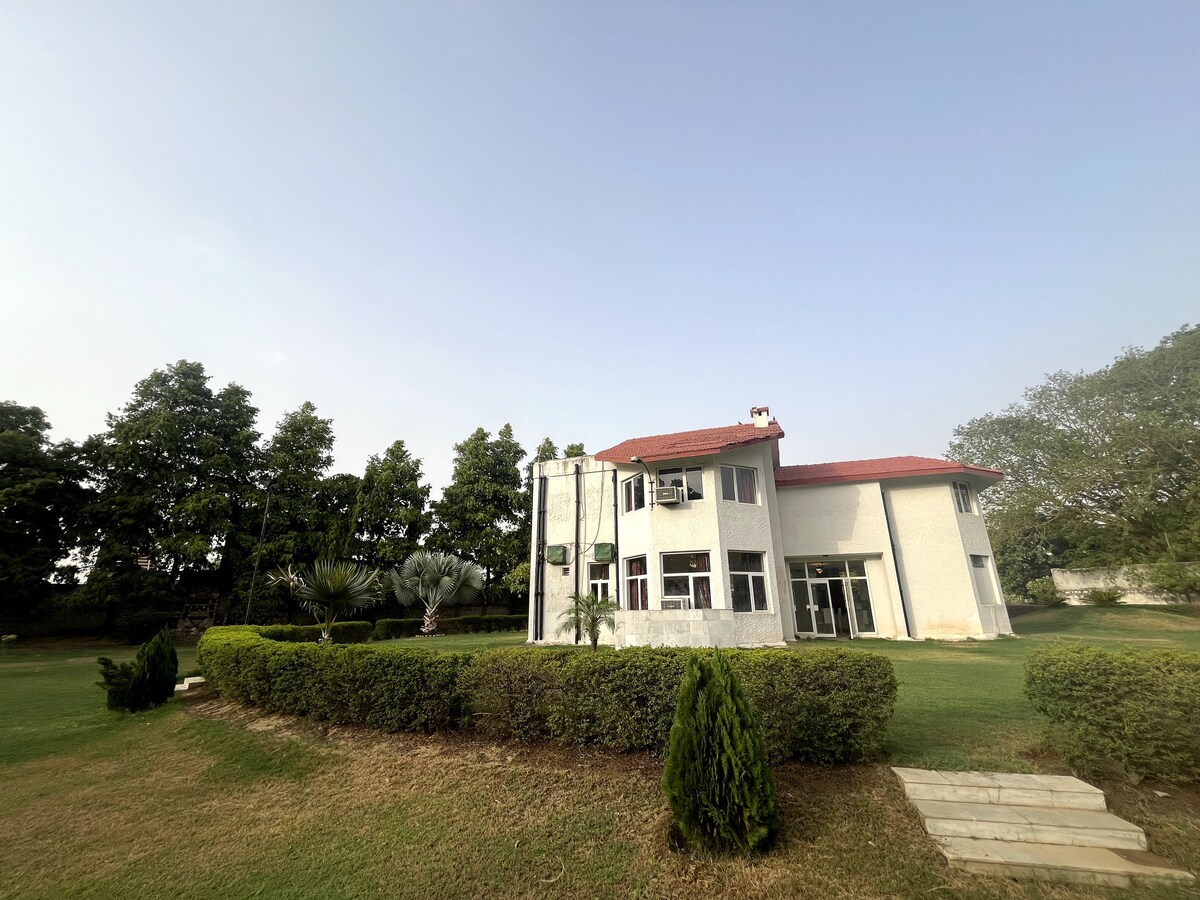 A spacious farmhouse is situated on a green landscape. The white structure features large windows and a red roof, surrounded by landscaped hedges and trees. The expansive lawn offers a serene environment, with a clear sky overhead enhancing the open space.