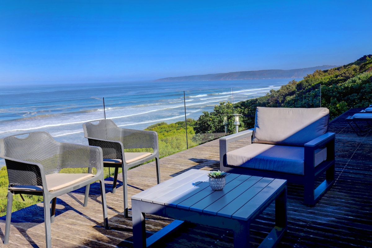 A deck area is furnished with a table and two armchairs, offering expansive views of rolling waves and a blue sky. Lush greenery is visible in the foreground, adding a natural touch to the outdoor setting.