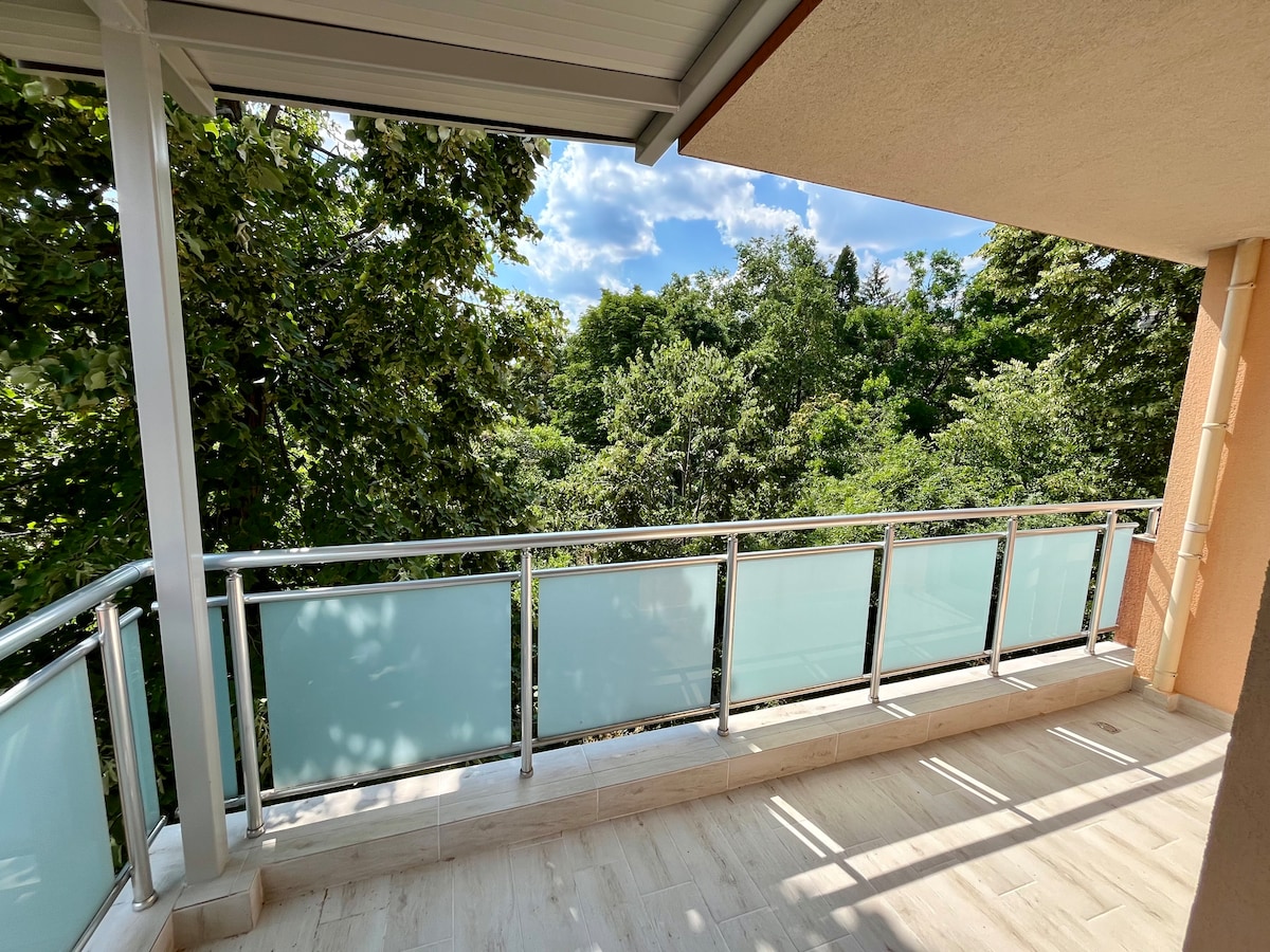 A spacious terrace is surrounded by lush greenery, offering a peaceful view of trees under a bright blue sky. The area features a glass railing that enhances the open feeling, and the flooring is designed with light-colored tiles.