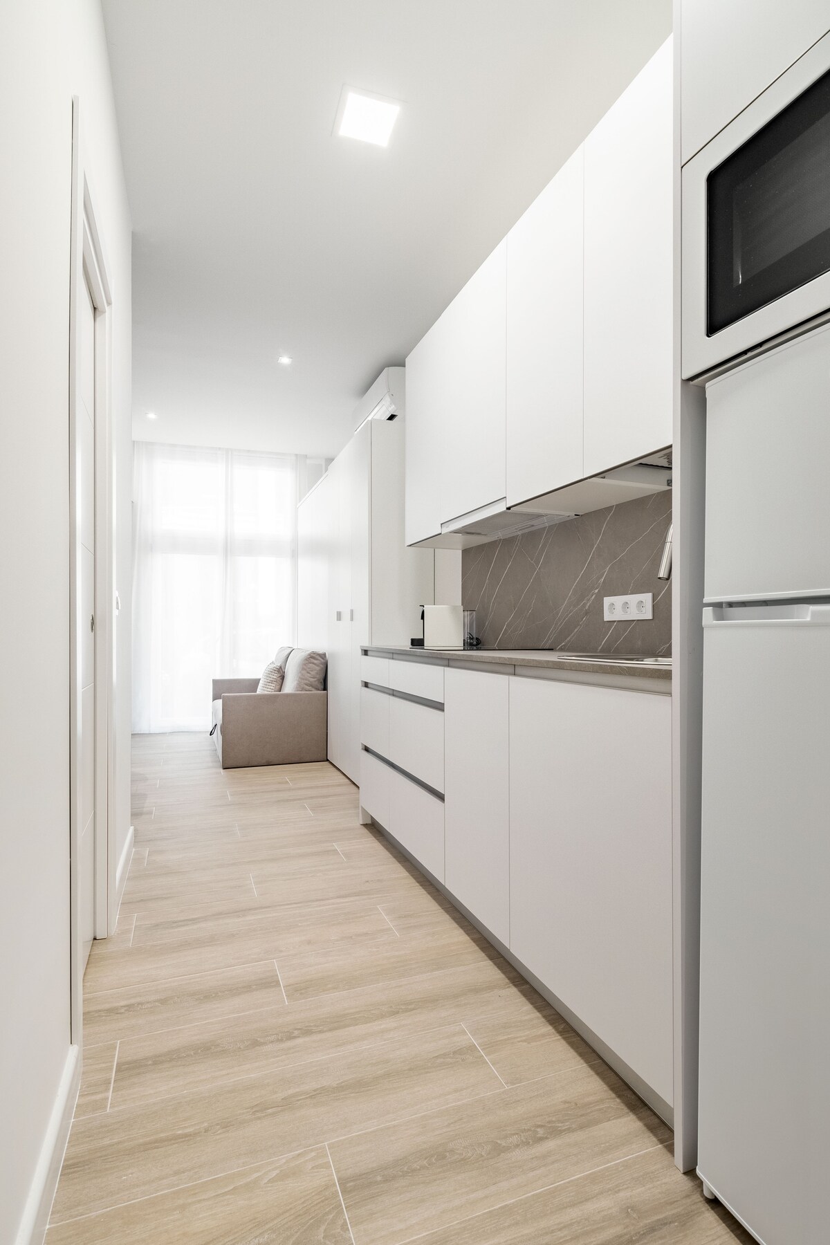 A sleek, modern kitchen is depicted, with white cabinetry and minimalist countertops. Light wood flooring extends throughout the space. Natural light filters through large windows into the adjoining area, where a sofa is partially visible, contributing to the open and airy environment.