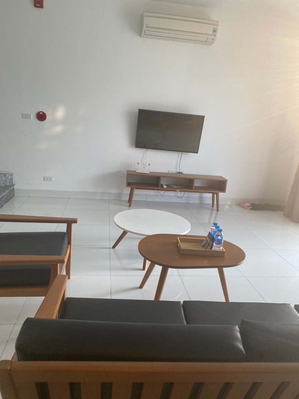 A modern living area is highlighted by a sleek television mounted on the wall. A light wood coffee table sits in front of a comfortable seating arrangement, which includes a dark couch and a single armchair. Natural light fills the space, creating an airy ambiance.