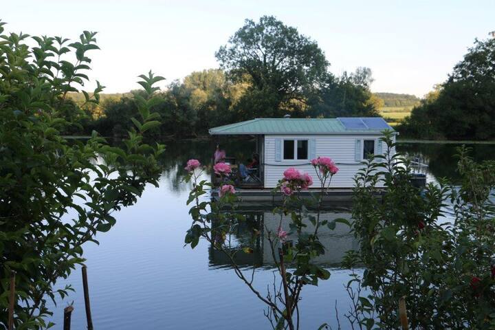 A floating cottage is observed on tranquil waters, surrounded by lush greenery and blooming pink flowers. The structure features a porch area, reflecting a serene escape into nature. The calm lake adds to the peaceful atmosphere of this unique getaway.