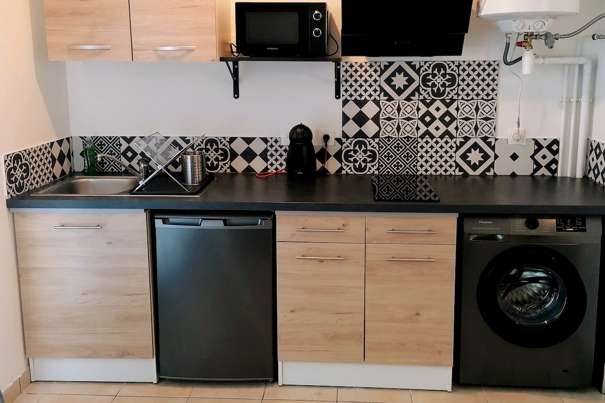 A modern kitchen area is displayed, featuring sleek wooden cabinetry and a dark countertop. A stainless steel sink is positioned next to a built-in dishwasher and washing machine. Decorative black and white tiles adorn the wall, complemented by a microwave and coffee maker on the shelf above.