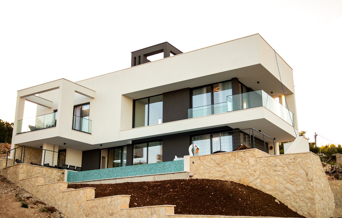 The modern villa is set against a hillside, featuring sleek lines and large glass windows. A spacious balcony encircles the upper floor, and a tranquil blue pool is visible at the ground level. The landscaped area includes terraced stonework for added visual interest.