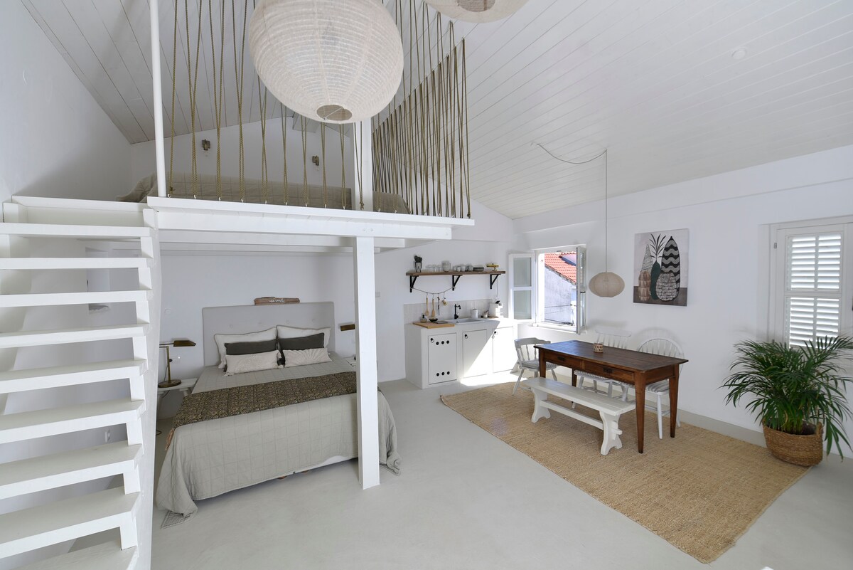 A bright and airy room features a spacious bed layered with neutral bedding, complemented by delicate pendant lights above. A dining area with a wooden table sits nearby, with a large potted plant adding a touch of greenery. Large windows provide ample natural light.