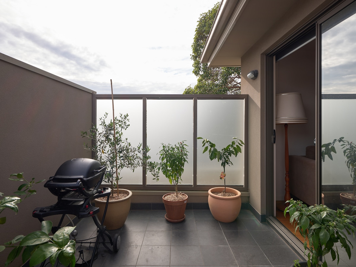 A spacious balcony is shown, featuring a barbecue grill beside two potted plants. Soft natural light fills the area, with a partially sunlit wall offering a gentle contrast. The glass railing provides a sense of openness while maintaining privacy.