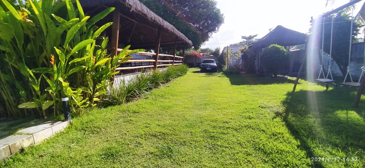 A spacious green lawn is framed by vibrant foliage and tall trees. A shaded area with a thatched roof provides outdoor seating options. A driveway runs through the garden, leading to a parked car, with sunlight filtering gently across the landscape.