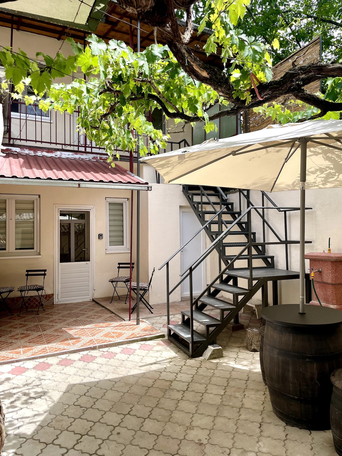 An outdoor courtyard is featured, shaded by a large grapevine. A spiral staircase leads to an upper level, while two small bistro tables and chairs are arranged on the tile flooring. A grill area is visible, accompanied by decorative barrels.