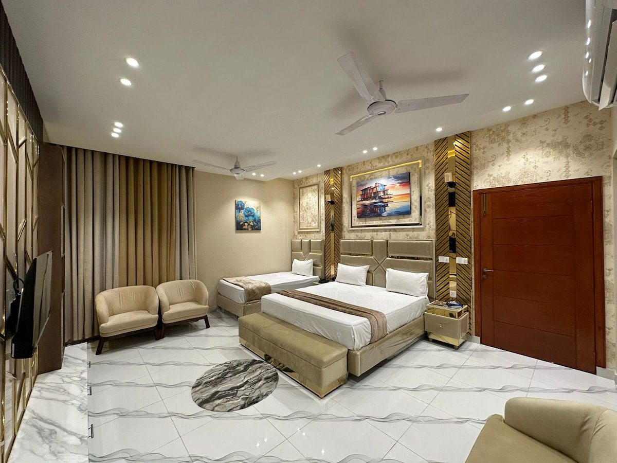 A spacious hotel room features a large bed with fresh linens, complemented by a cozy seating area with two light-colored chairs. Modern artwork adorns the walls, and natural light filters through elegant curtains. A sleek television is mounted on a stylish wall unit.