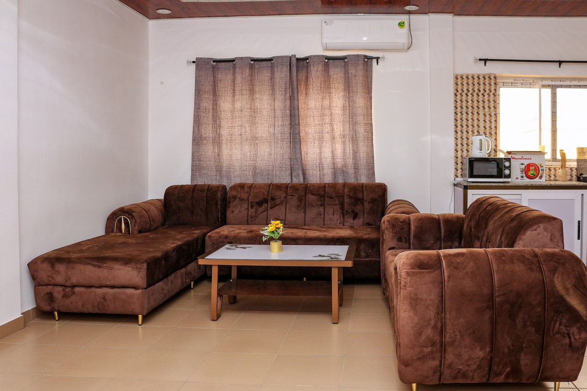 A spacious living area is furnished with a large brown sectional sofa and a low coffee table. Natural light enters through a window covered by dark curtains, while a small flower arrangement adds a subtle touch to the table.
