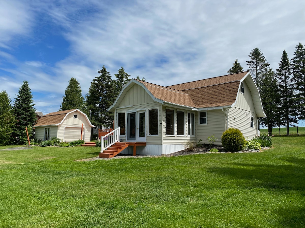 A light-colored house with a spacious porch is surrounded by lush green grass and trees. The well-maintained lawn leads to a second building, possibly a garage. The sky above is partly cloudy, providing a bright and inviting atmosphere.