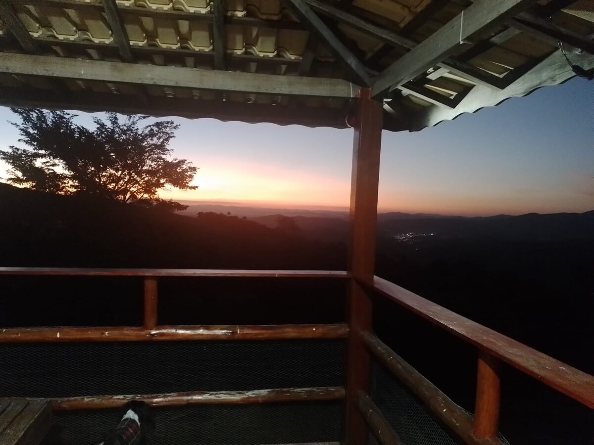 A wooden balcony offers a scenic view of the horizon during sunset, with muted colors blending in the sky. The surrounding landscape features hills and trees, creating a peaceful atmosphere for relaxation and contemplation.