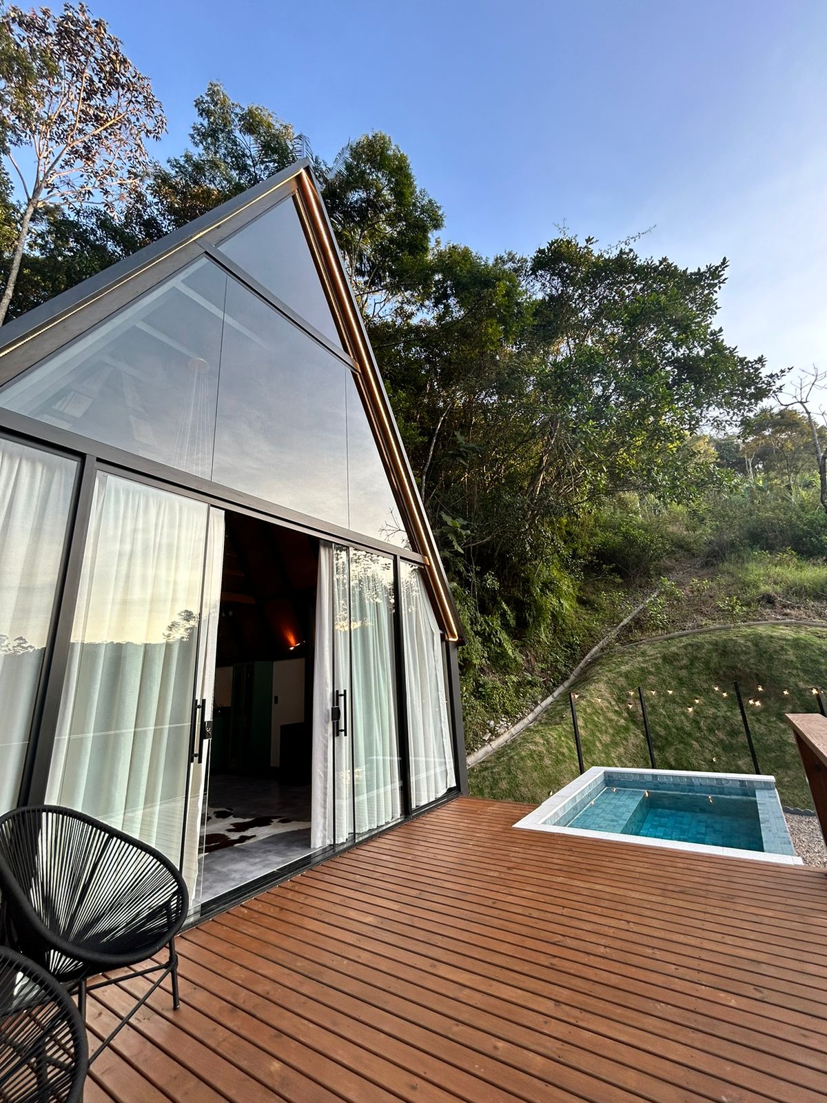 A modern cabin features large glass walls that open to a wooden deck. An inviting plunge pool is visible next to the deck, surrounded by greenery. The tranquil setting offers a seamless integration of indoor and outdoor living.