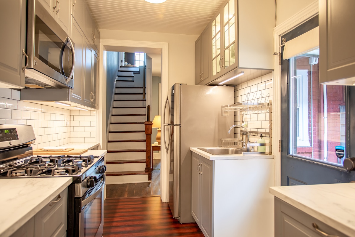 The kitchen features modern stainless steel appliances, including a gas stove and refrigerator. White cabinetry and countertops are complemented by a tiled backsplash. Natural light is provided by a door leading to the outside, while a staircase to the living area is visible at the end of the room.