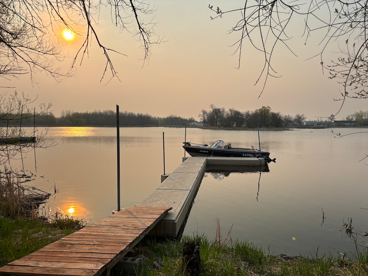 A private dock extends into calm waters, with a boat moored alongside. The sun rises softly, casting a warm glow on the surface. Trees frame the scene, reflecting hints of green, while the tranquility of the setting is evident in the stillness of the water.