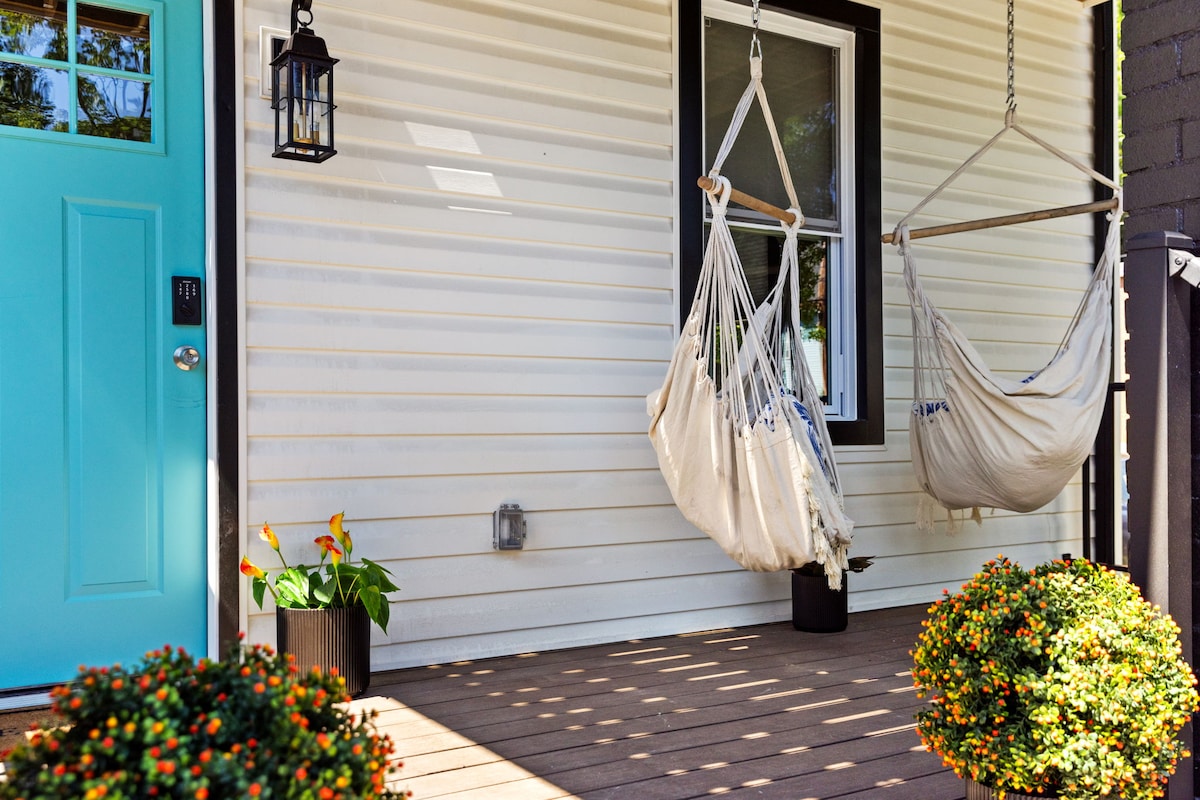 A front porch is showcased with two hammock swings suspended from the ceiling. A bright blue door is visible, framed by a lantern and a potted plant. Small flowering plants in black and orange pots add a touch of greenery to the space.
