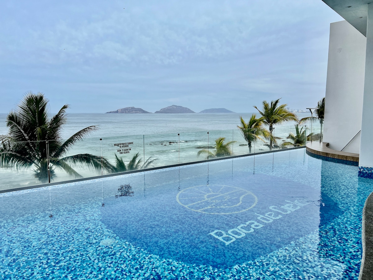 A serene infinity pool is positioned at the edge of the property, offering unobstructed views of the ocean and distant islands. Clear blue waters reflect the sky, while palm trees frame the scene, enhancing the coastal ambiance.