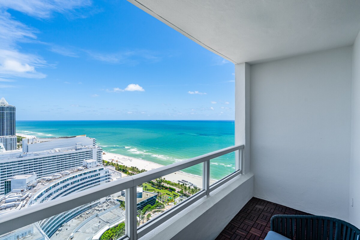 A private balcony offers sweeping views of the turquoise ocean and Miami Beach coastline. The scene captures vibrant blue waters meeting a clear sky, with the city skyline in the foreground, creating a serene and inviting outdoor space suitable for relaxation.