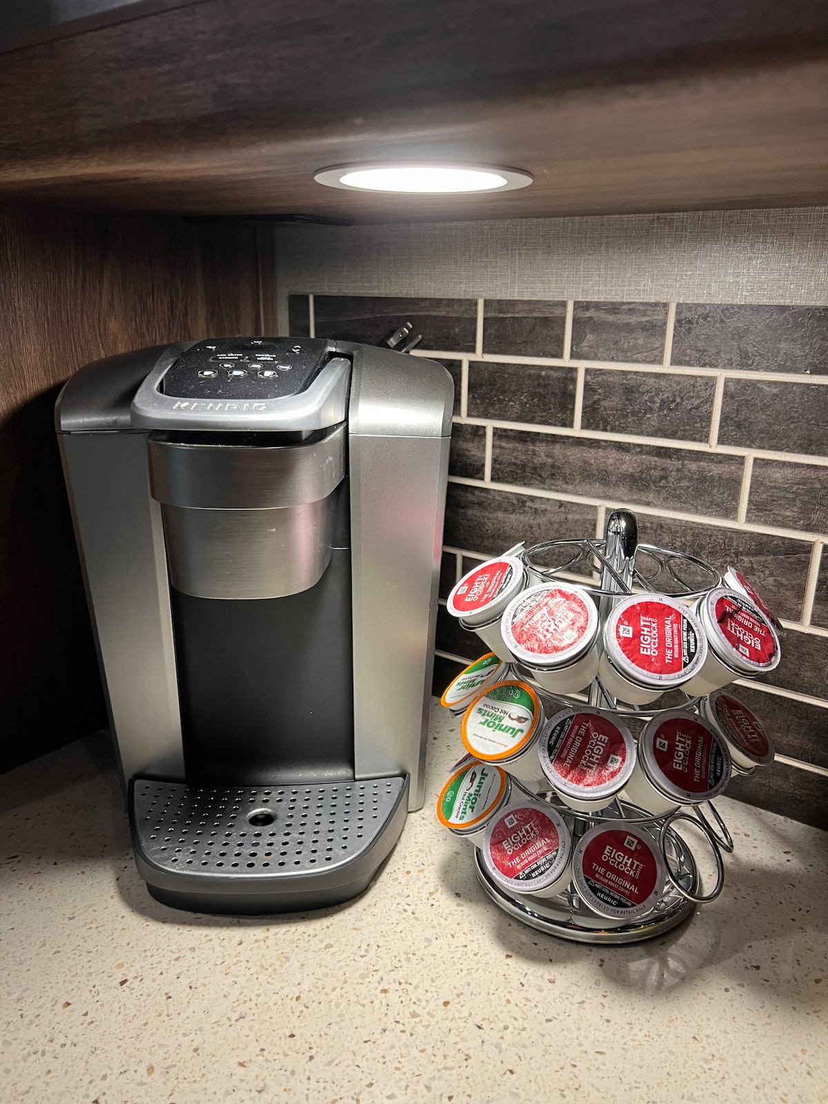 A coffee maker is positioned on a countertop beneath soft under-cabinet lighting. Next to it, a circular rack displays a variety of coffee pods in colorful packaging, offering options for a morning brew.