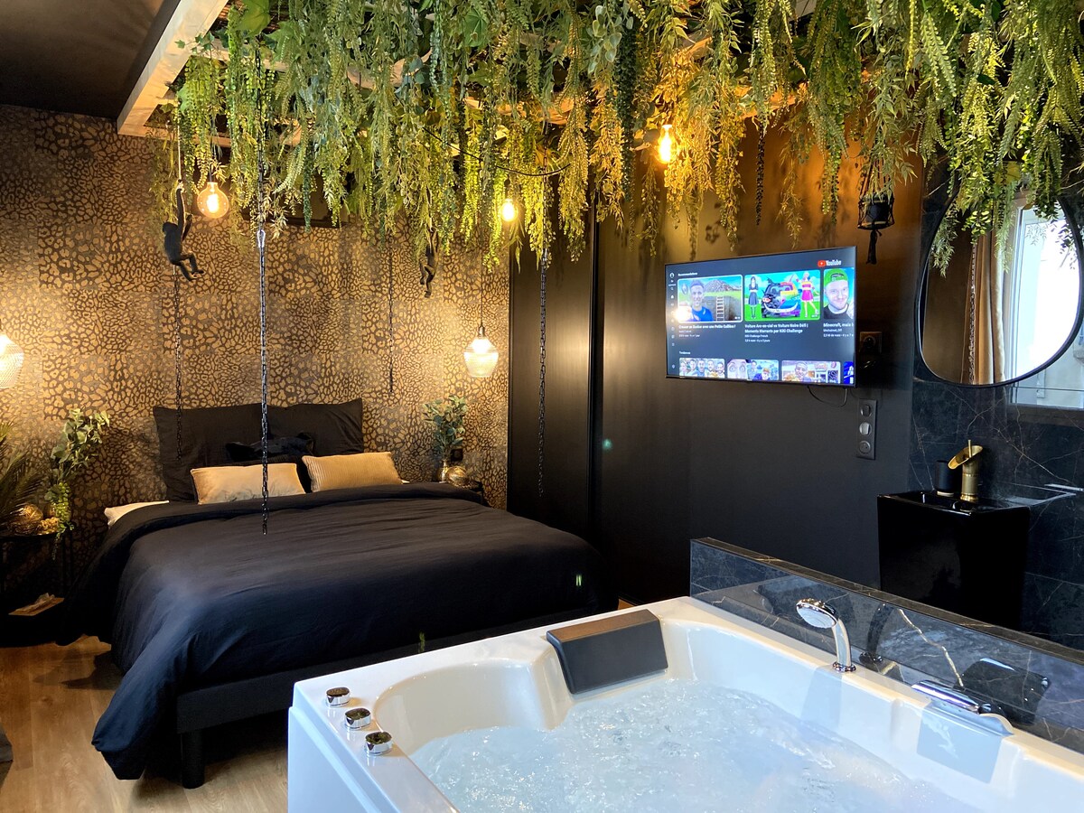 An intimate space features a spacious bed dressed in dark linens, with ambient lighting provided by pendant lights. A large therapeutic bathtub is visible in the foreground, complemented by greenery adorning the ceiling. A mounted TV is positioned on the wall opposite the bed.