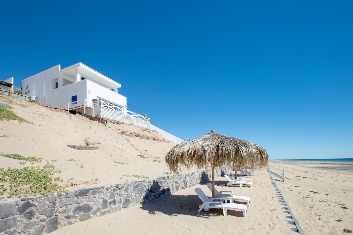 A beachfront property is positioned slightly elevated from the sandy shore, featuring a thatched umbrella providing shade for two lounge chairs. The coastline extends toward a clear blue horizon under a bright sky, emphasizing the serene atmosphere of the secluded location.