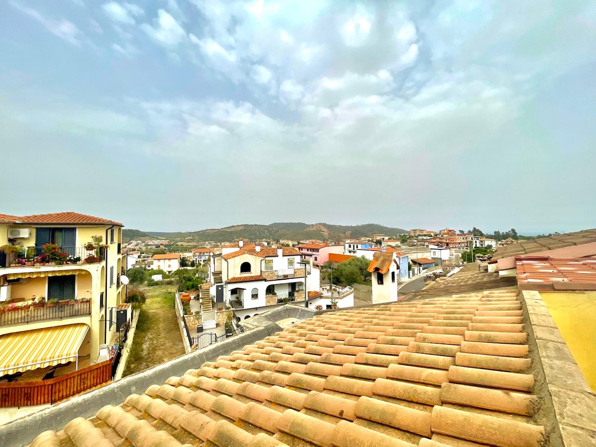 A panoramic view captures a residential area featuring low-lying houses with terracotta roofs. Rolling hills are visible in the distance under a cloudy sky, creating a subdued atmosphere. The scene suggests a quiet, suburban environment with various architectural styles.