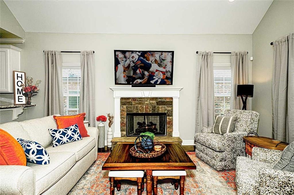 A welcoming living room features a comfortable sofa with patterned cushions, paired with an armchair and a coffee table. A stone fireplace serves as a focal point, and a television is mounted above it. Soft natural light filters through the neatly draped curtains.