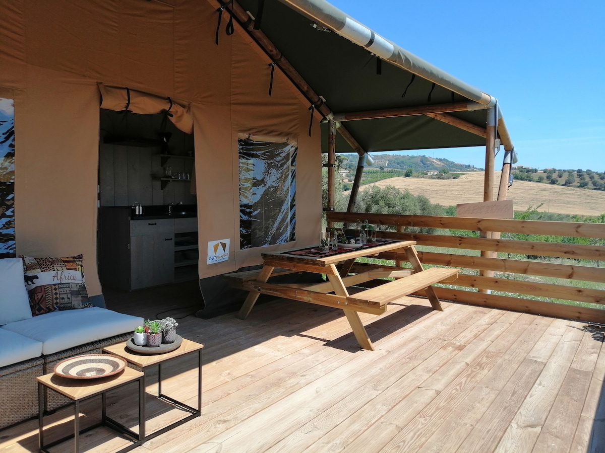 A spacious glamping tent is set against a scenic landscape, with natural light filtering through large windows. A wooden deck features a picnic table, surrounded by greenery, and an outdoor seating area with a small coffee table. The inviting atmosphere showcases comfortable furnishings.