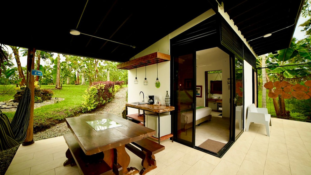 A charming outdoor dining area is visible, featuring a wooden table and benches. A kitchenette area is included with overhead pendant lighting. Large glass doors lead into a spacious interior, framed by lush greenery and a pathway that meanders through the garden.