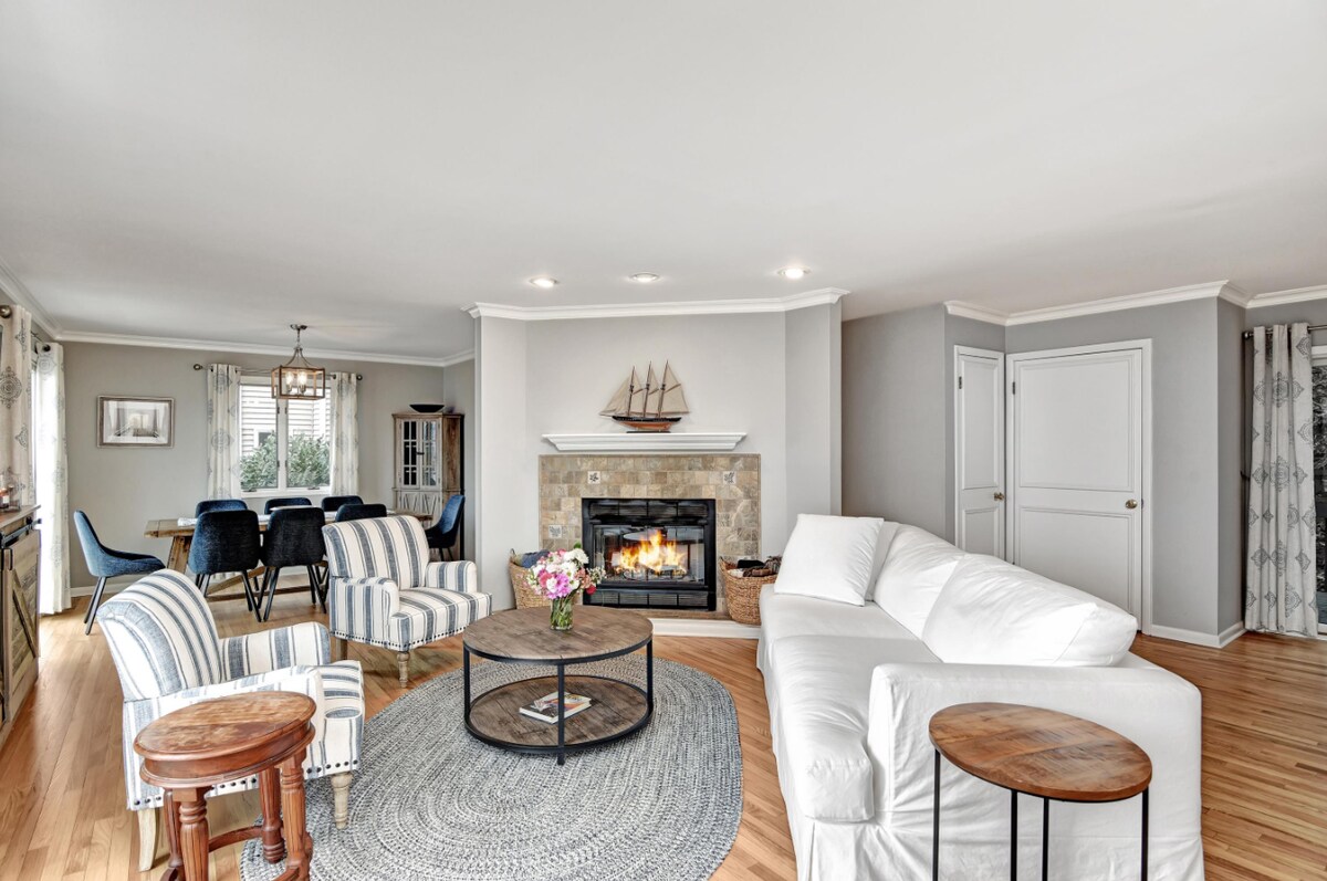 An inviting living area features a cozy white sectional sofa paired with striped armchairs. A round coffee table sits atop a soft area rug, and a stone fireplace provides a warm focal point. Natural light filters through large windows, highlighting the open space.