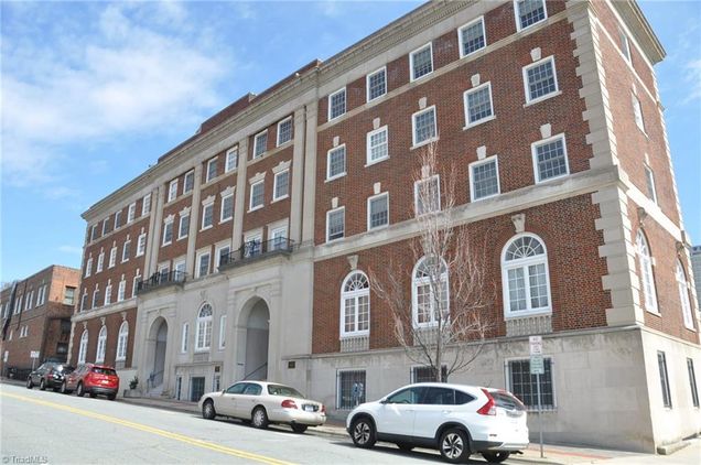 A four-story brick building is presented, featuring a classic architectural style. Large arched windows dominate the façade, with smaller rectangular windows situated above. A mixture of vehicles is parked at the curb, and a clear blue sky complements the scene.