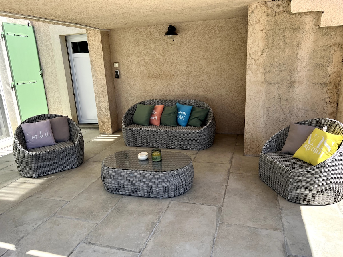 An outdoor seating area is presented with four woven chairs arranged around a low coffee table. Colorful cushions are placed on the chairs, adding a touch of comfort. The textured walls and tiled floor create a relaxed atmosphere.