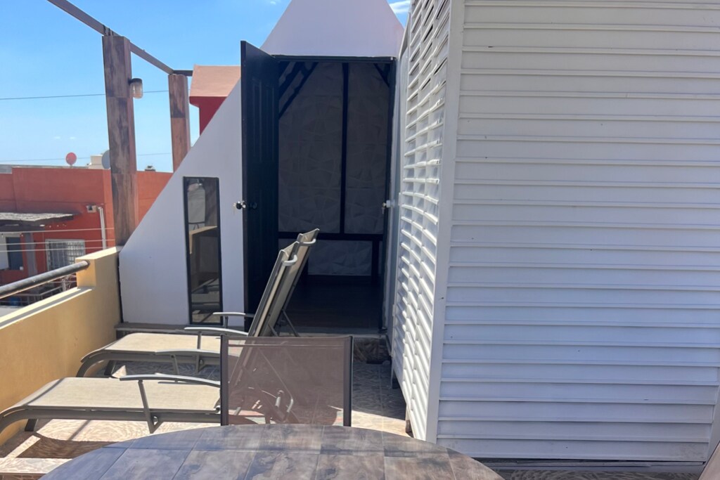 A small outdoor area features a table and several lounge chairs arranged on a wooden deck. An entrance leads to an enclosed space, partially visible, with a contrasting colored exterior. Bright sunlight illuminates the setting, creating a relaxed atmosphere.