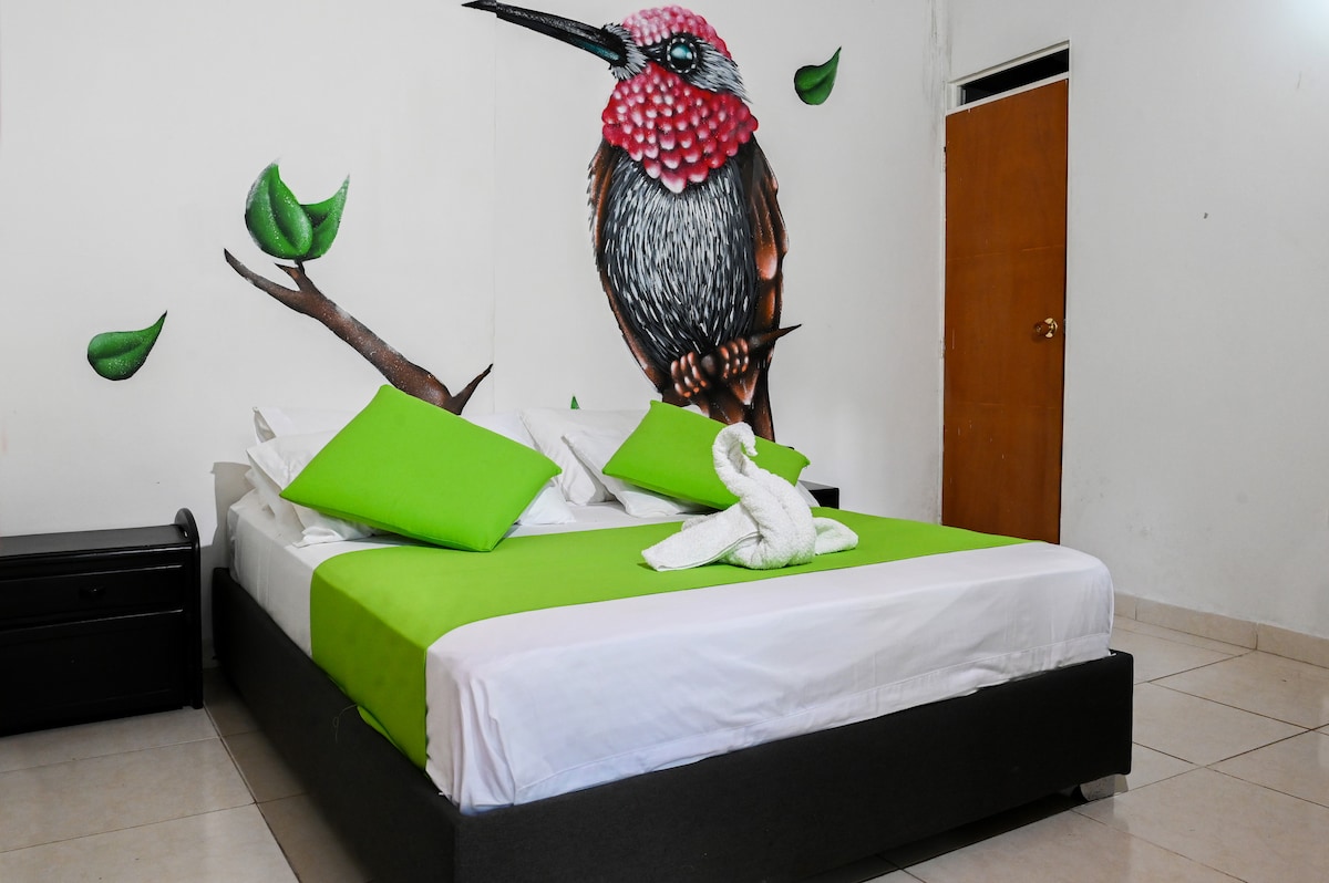 A spacious sleeping area features a large bed with bright green accent pillows. A colorful mural of a bird on a branch adorns the wall, complemented by fresh green leaf details. A wooden drawer unit is positioned against the opposite wall, and natural light enters through a nearby door.