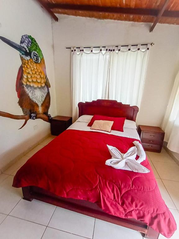 The room features a wooden bed with a bright red quilt and white pillowcases. Nightstands are positioned on each side of the bed. A large mural of a colorful hummingbird adorns one wall, while light filters through white curtains covering a window.