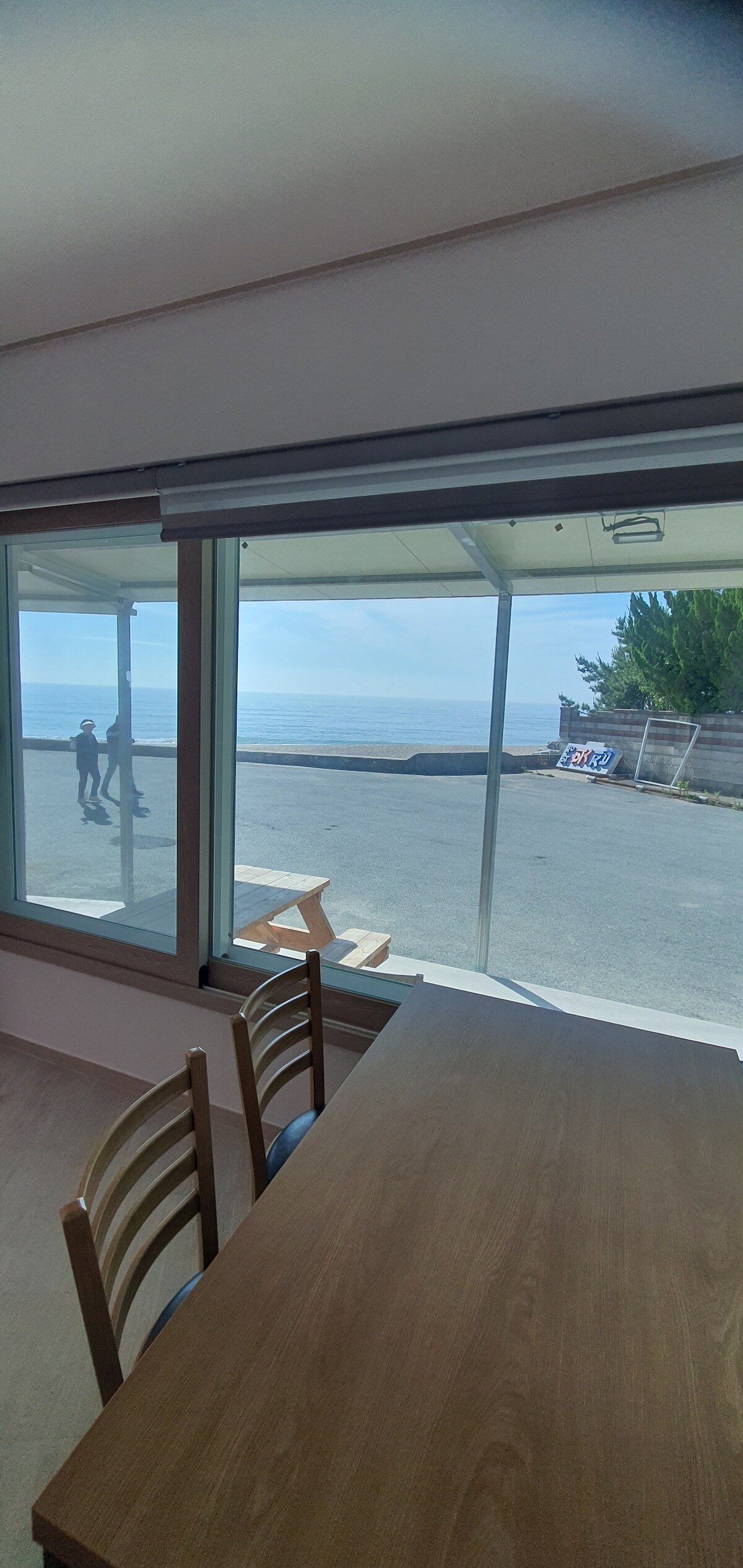 A calm view of the ocean is framed by large glass windows. Natural light floods the interior space, where a wooden table with a simple design is positioned. Outside, a pathway and greenery are visible, adding to the serene backdrop.