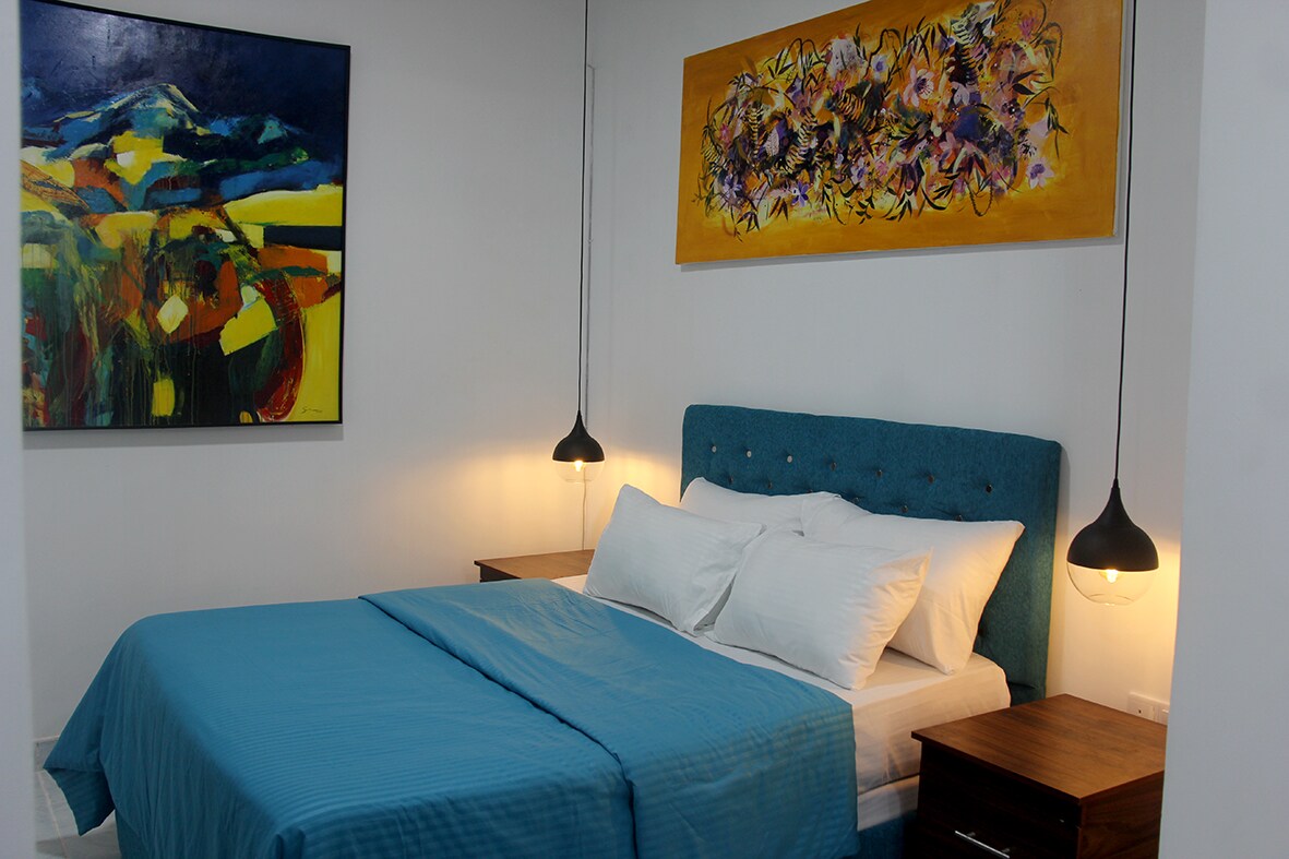 A well-appointed sleeping area features a spacious bed adorned with a textured blue blanket. Above the bed, a vibrant artwork adds color to the room. Pendant lighting casts a soft glow, complemented by dark wooden nightstands on either side.