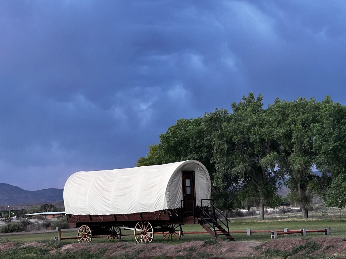 A vintage wagon is positioned on a grassy area, with large trees and distant mountains visible in the background. The wagon features a rounded canvas top and wooden wheels, framed by an expansive sky filled with dramatic clouds.