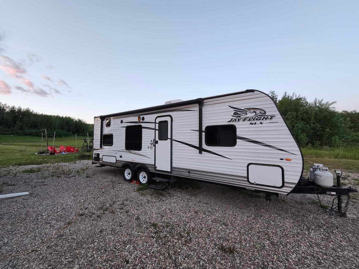 A parked camper is shown featuring a white exterior with black accents. The landscape includes a grassy area and a red seating arrangement in the background. A gravel surface is visible beneath the camper, with trees lining the distance.