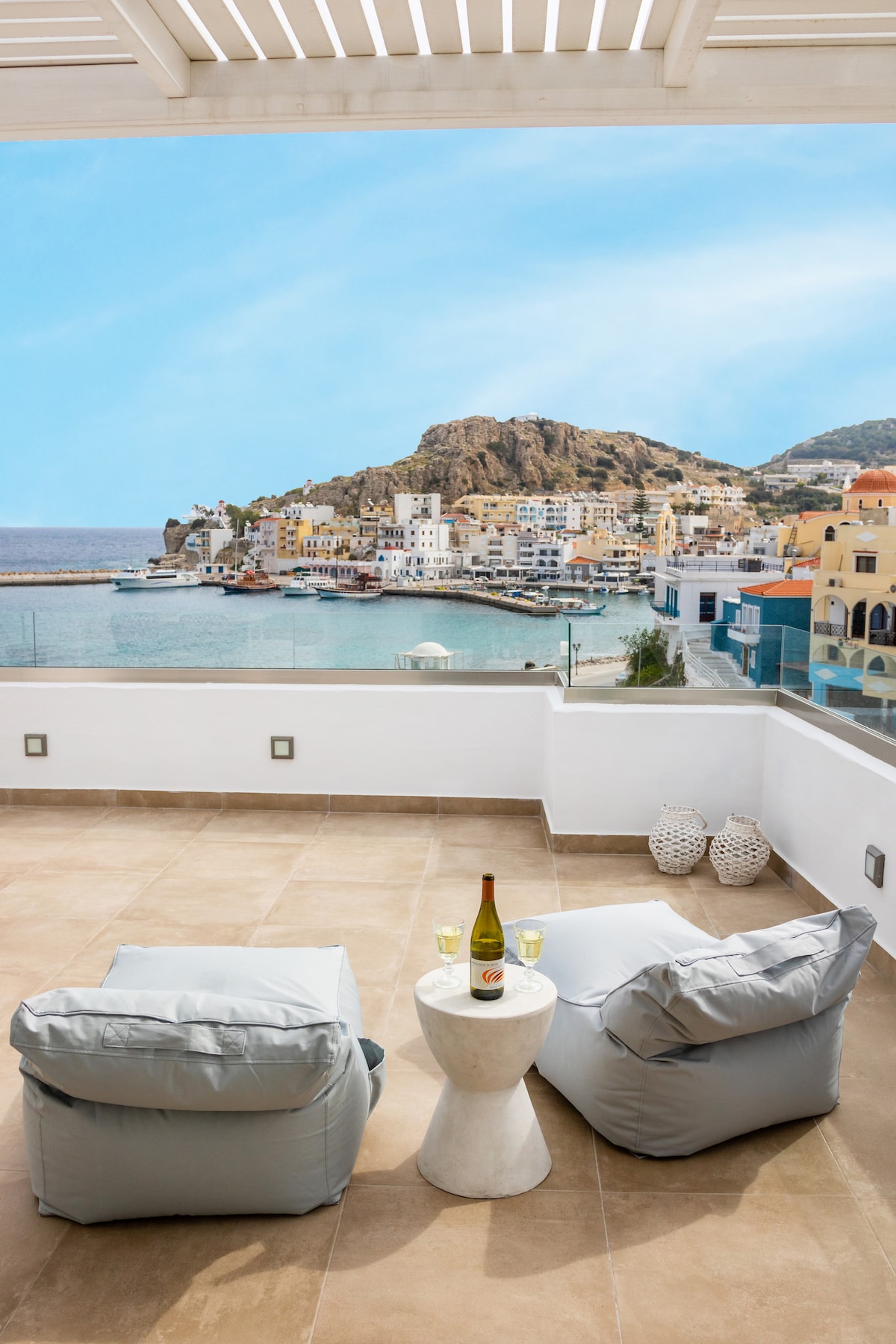 A spacious terrace is presented, featuring two soft seating areas and a small white table. A bottle of champagne sits on the table, accompanied by two glasses. The stunning panoramic view showcases the Aegean Sea and the colorful buildings of Pigadia in the background.