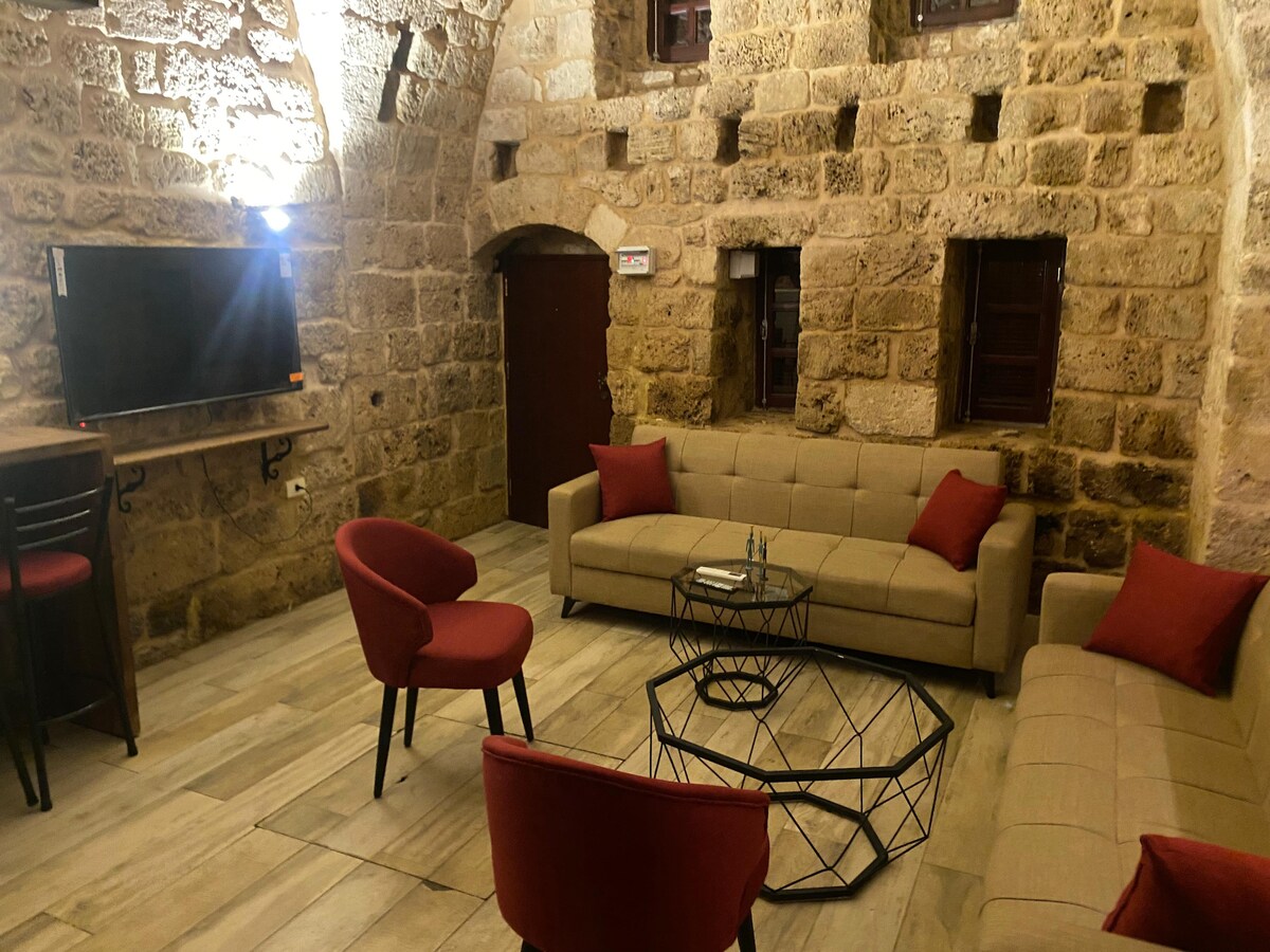 A spacious living area showcases exposed stone walls and wooden windows. Two comfortable couches in neutral tones, accented with red cushions, are arranged around a circular glass table. A smart TV mounted on the wall provides entertainment, complementing the inviting ambiance.