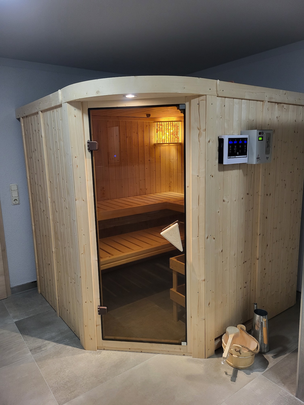 A wooden sauna is featured with a glass door, allowing visibility into the interior. Soft lighting highlights the natural wood benching inside. A digital control panel is visible outside, alongside a wooden bucket and ladle positioned on the floor.