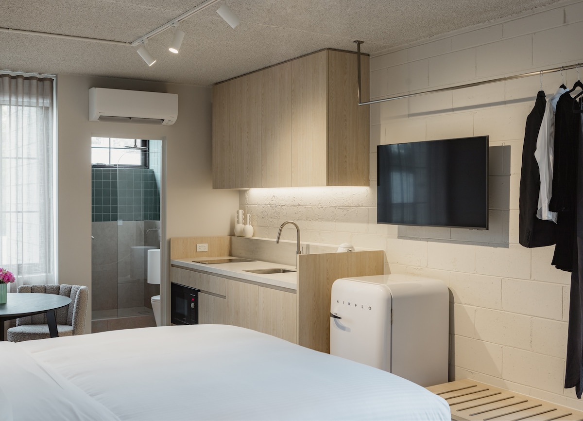 A modern studio apartment is presented with an open layout. A sleeping area features a spacious bed, while a kitchenette is equipped with essential appliances. A small dining table is included in the corner, and a television is mounted on the wall. A bathroom is visible, showcasing a walk-in shower.