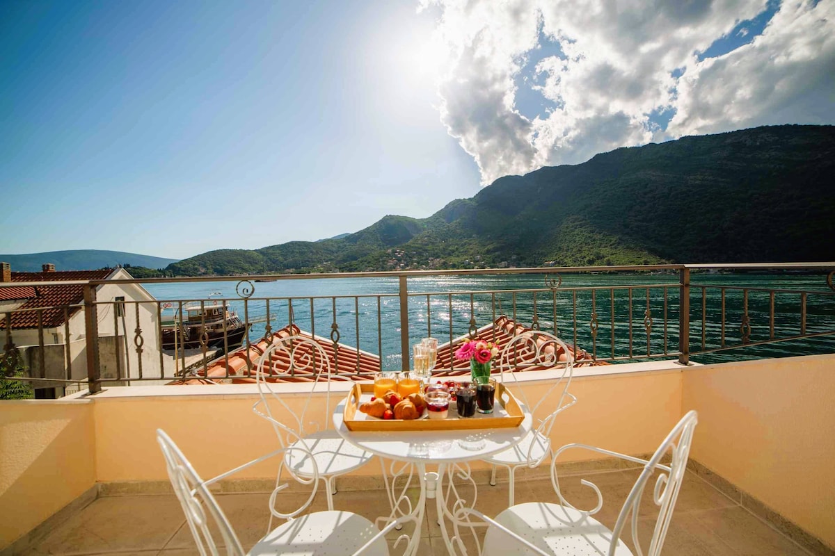 A private terrace presents a round table and four white, wrought-iron chairs, adorned with a vibrant spread of fruits and beverages. Boka Bay's shimmering waters and verdant mountains serve as a scenic backdrop under a bright sky.