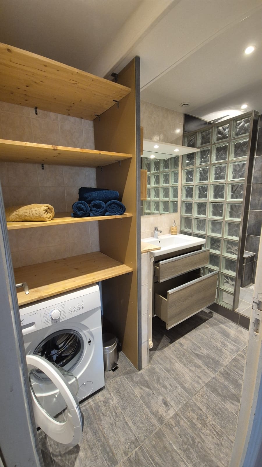 A compact bathroom features a glass block shower and a modern sink with storage beneath. Shelves hold neatly rolled towels in blue. A washing machine is positioned at the bottom of the space, while light from above accentuates the functional arrangement.