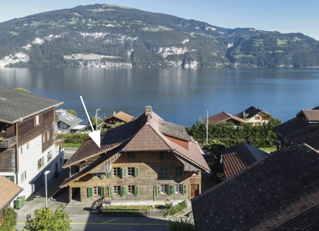 An aerial view of a historic wooden building situated near the Thunersee. Surrounding mountains provide a scenic backdrop. The structure features a sloped roof and large windows, with greenery framing the property. Nearby homes create a quaint village atmosphere.