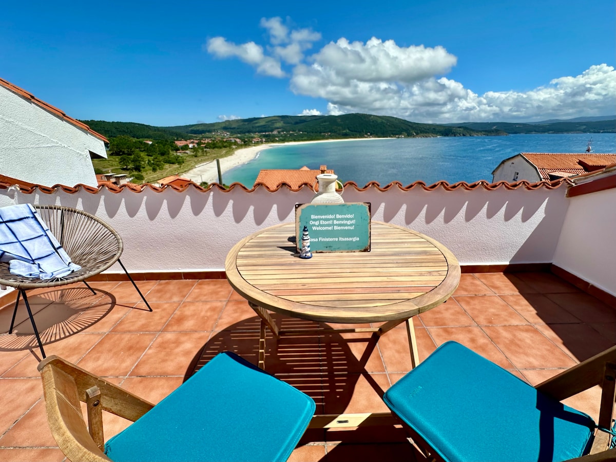 A terrace is shown overlooking the coastline, featuring a wooden table with two teal chairs. A relaxing atmosphere is created by the scenic views of the beach and ocean under a bright sky, complemented by a gentle sea breeze.