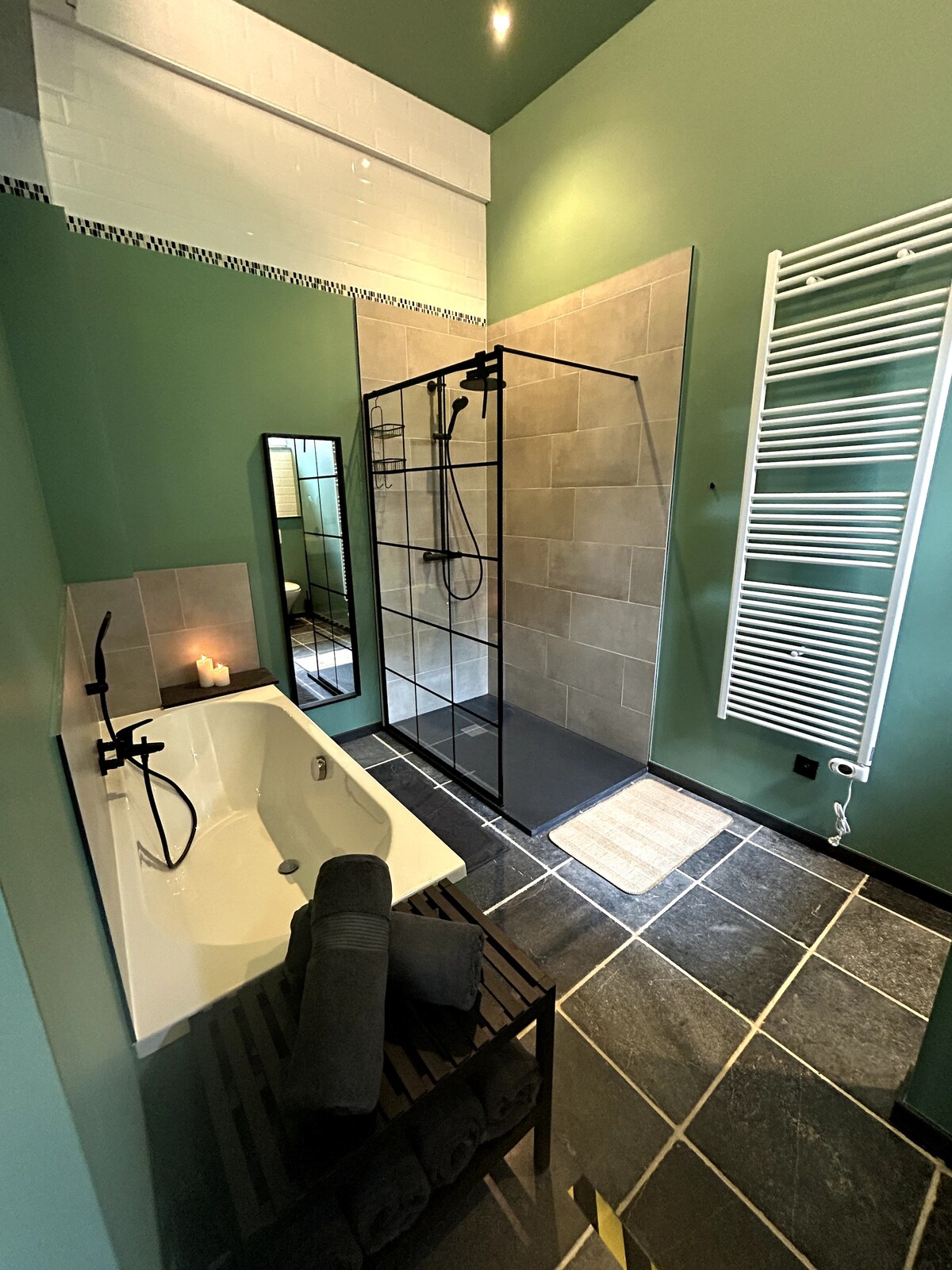 The bathroom features a modern design with green walls and stone tiles. A spacious bathtub and a walk-in shower are present, with a sleek glass partition. A full-length mirror reflects the space, alongside a white towel radiator mounted on the wall.